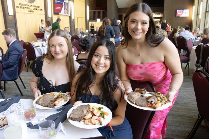 Senior Soiree Spring 2025 Two women sitting in chairs and one women standing to their right. All three are holding plates of food with beef and shirmp.