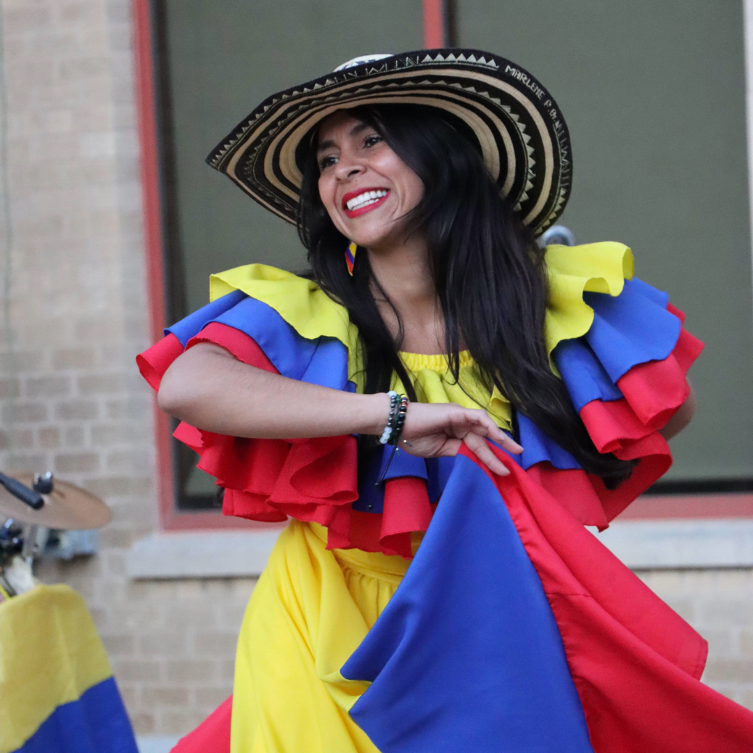woman with long dark hair wearing a hat in a colorful dress of red, blue, and yellow