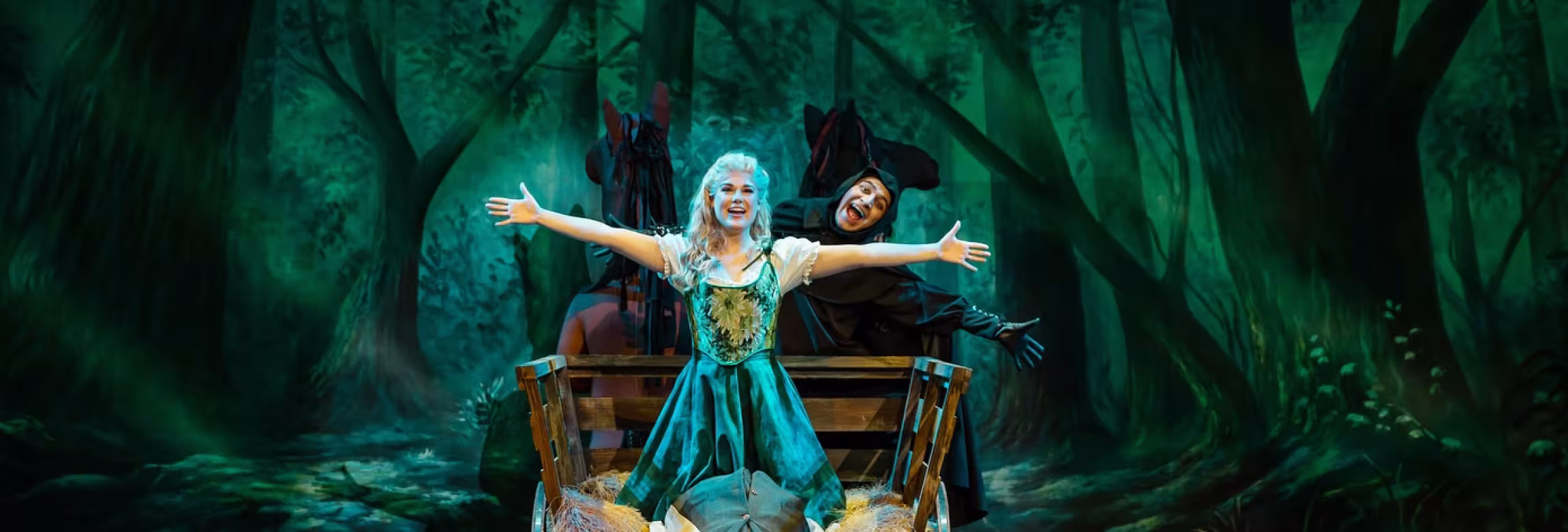 scene from a production, women in a cart with person in black behind. Both with arms spreadout and smiles.