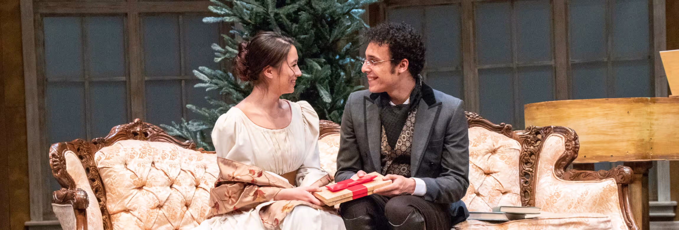 From a production, a women and man sitting on a couch holding a gift box.
