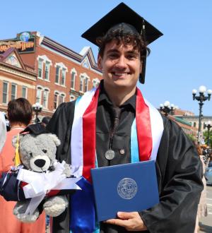 Man in a graduation cap and gown. He is holding a blue diploma and a teddy bear wearing a graduation cap Viterbo commencement